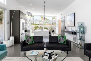 Ein modernes Wohnzimmer mit einer schwarzen Couch, grünen Kissen, einem Glastisch und einer Küche im Hintergrund.