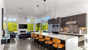A modern kitchen with a central island, bar stools, a flat-screen TV, and a large window overlooking greenery. - Modern Desert Oasis – Luxury Retreat (Palm Springs)