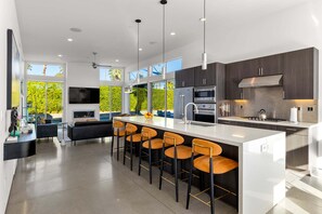 A modern kitchen with a central island, bar stools, a flat-screen TV, and a large window overlooking greenery. - Modern Desert Oasis – Luxury Retreat (Palm Springs)
