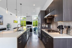 A modern kitchen with a central island, dark cabinetry, and a large window overlooking greenery. - Modern Desert Oasis – Luxury Retreat (Palm Springs)