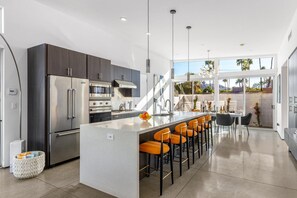 A modern kitchen with a central island, stainless steel appliances, and a bar area with orange chairs. - Modern Desert Oasis – Luxury Retreat (Palm Springs)