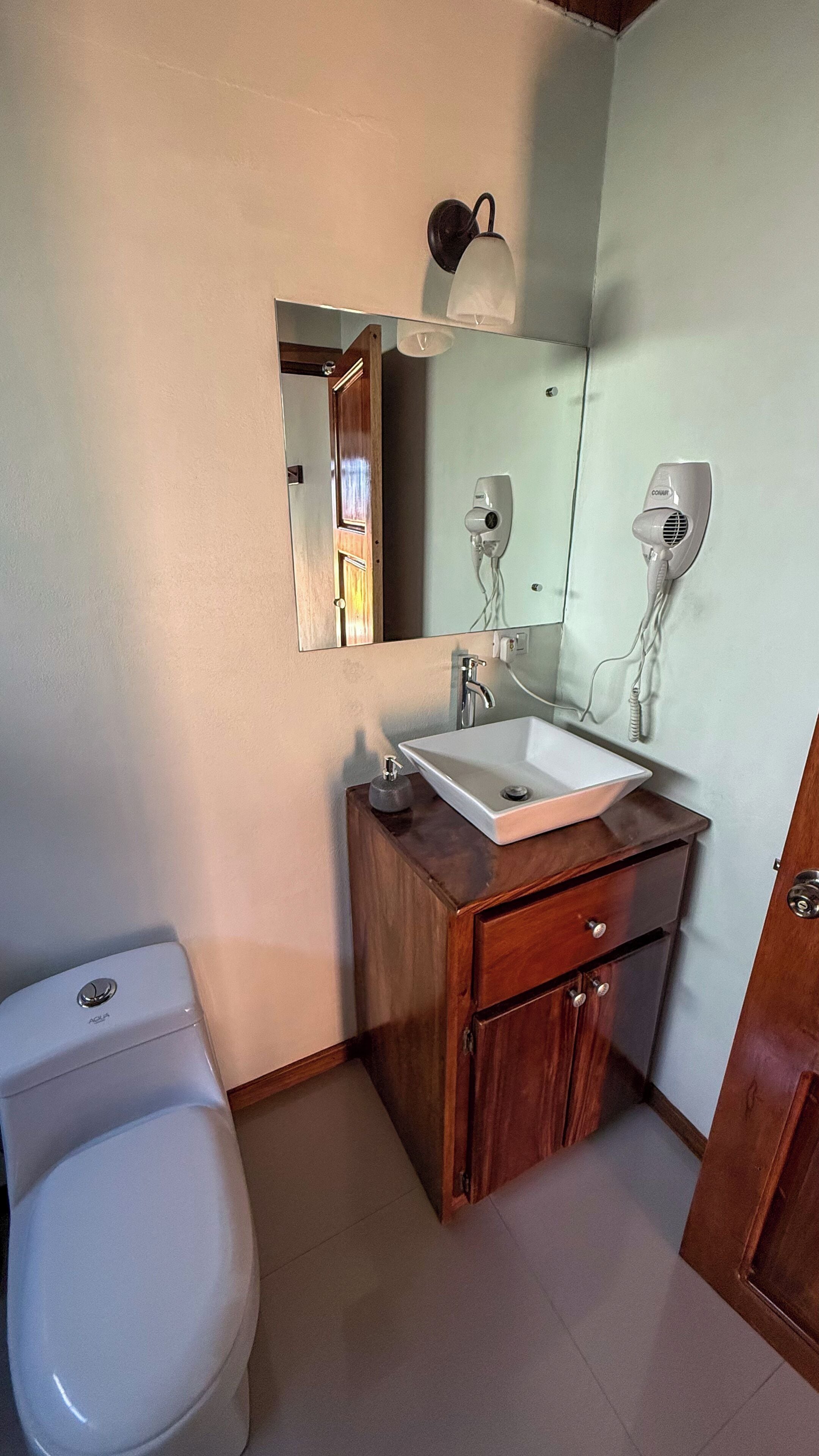 A bathroom with a white toilet, a wooden vanity with a sink, a mirror, and a hairdryer mounted on the wall.