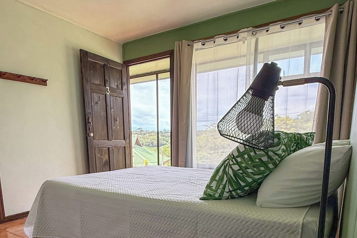 A room with a bed, a desk with a lamp, and a view of the outdoors.