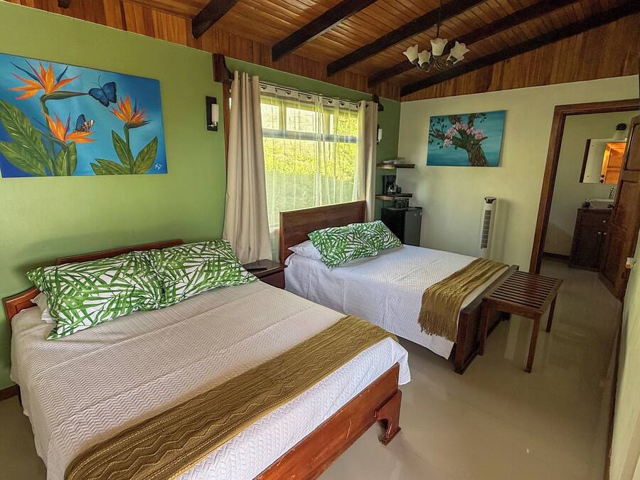 Habitación con dos camas, paredes verdes, vigas de madera en el techo y obras de arte con pájaros y flores.