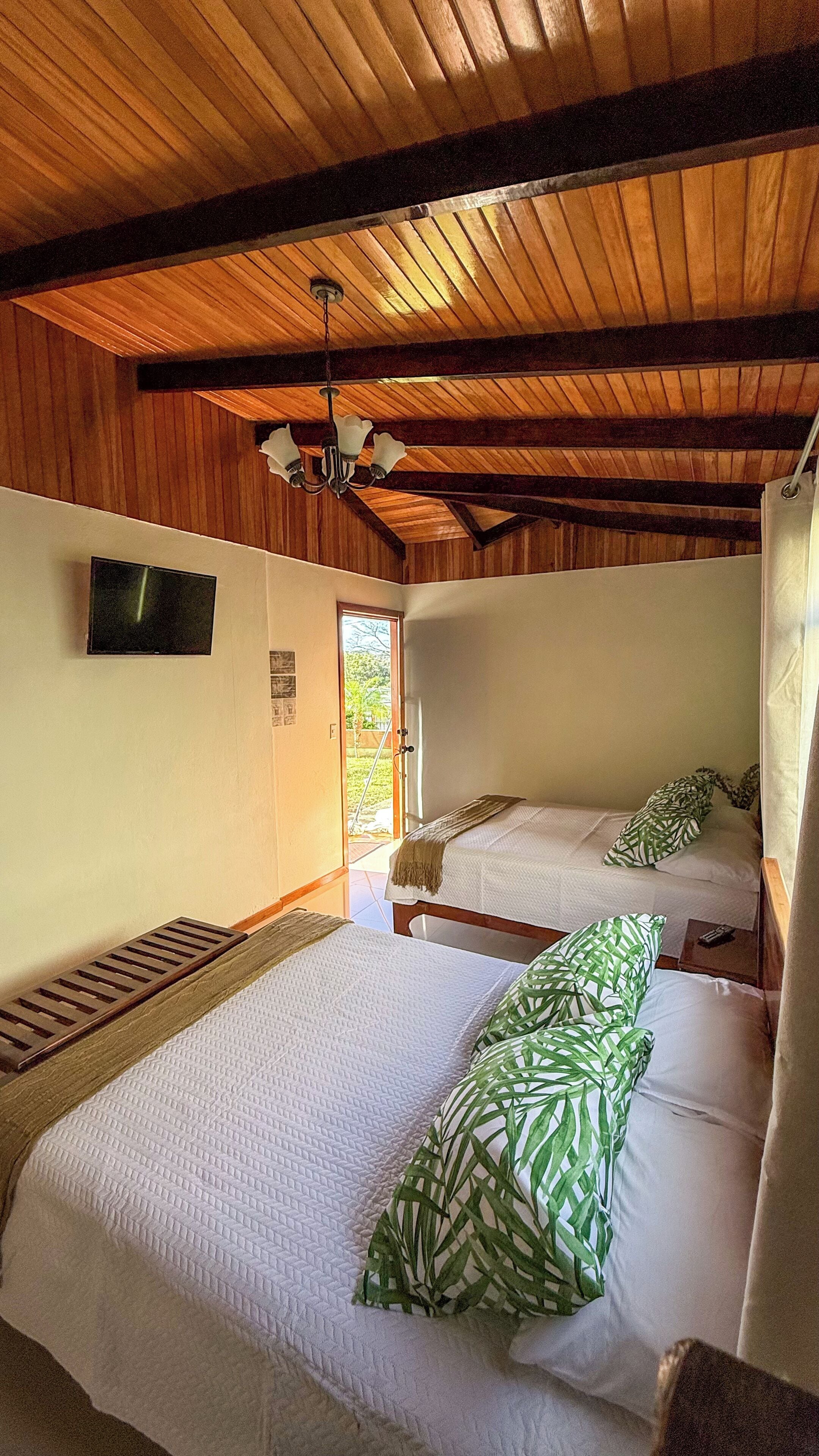 Ein Zimmer mit zwei Betten, Holzbalkendecke, einem an der Wand befestigten Fernseher und einer Tür, die nach draußen führt.
