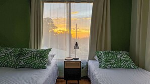 A room with two beds, each with green patterned pillows, a small table with a lamp, and a window with a view of a sunset. - Brumas de Valverde (Monteverde)