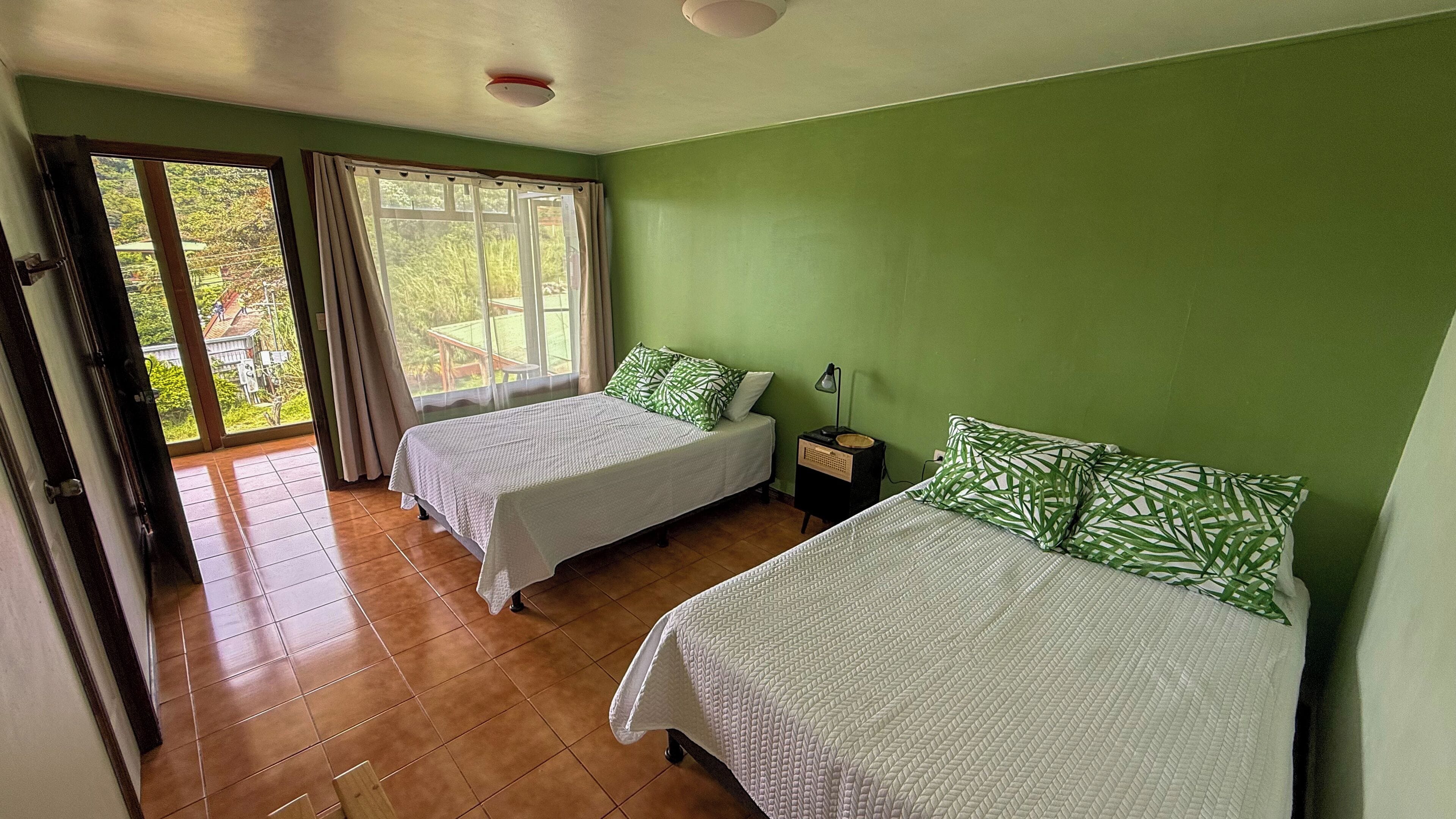 A room with two beds, a window with curtains, a nightstand with a lamp, and a view of greenery outside.