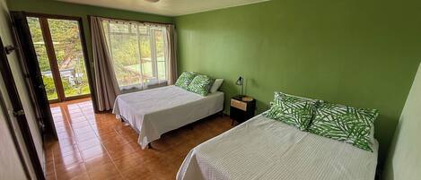 A room with two beds, a window with curtains, a nightstand with a lamp, and a view of greenery outside.