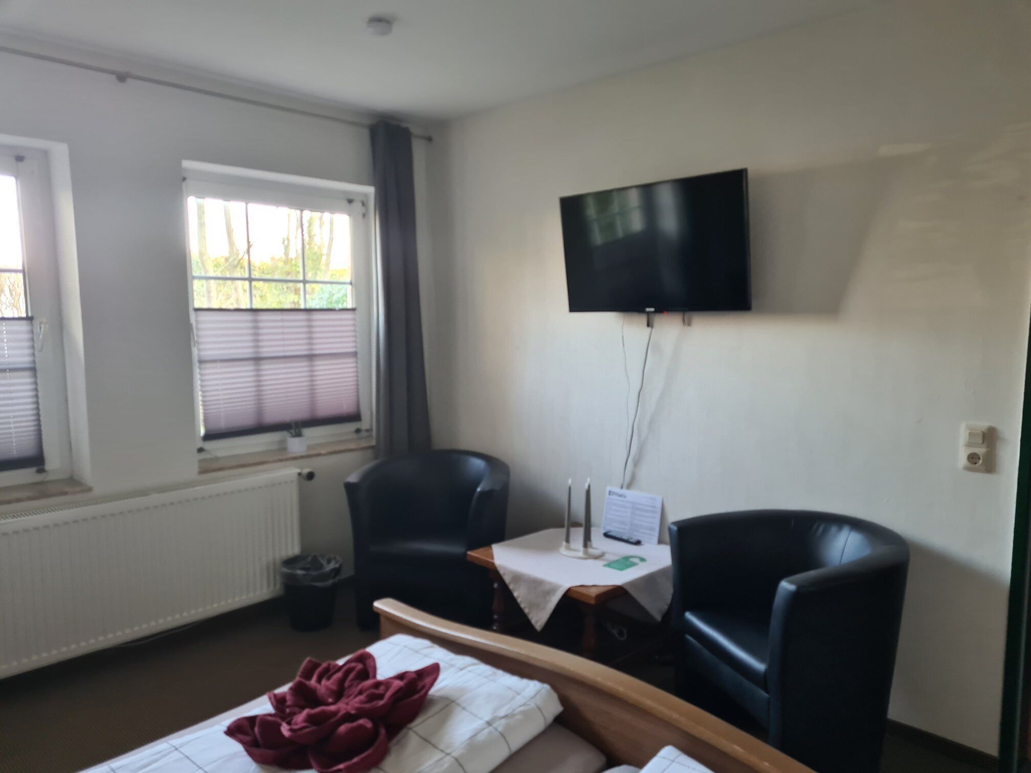 A hotel room with a bed, two chairs, a table with a lamp, a TV on the wall, and a window with blinds.