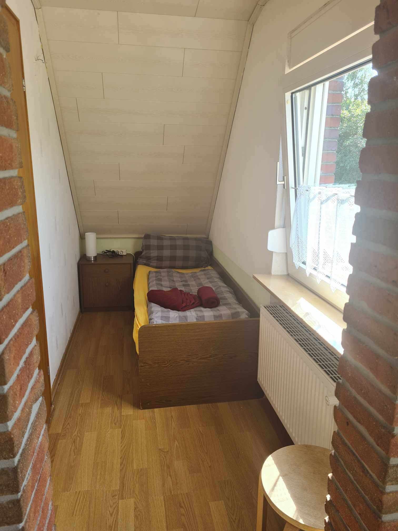 A small, sloped-roof bedroom with a single bed, a radiator, and a window with a view of the outside.