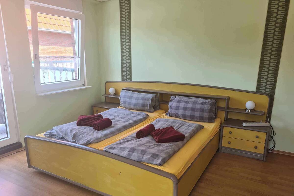 Una cama doble con dos almohadas y una colcha, una mesita de noche con lámpara, y una ventana con persianas.
