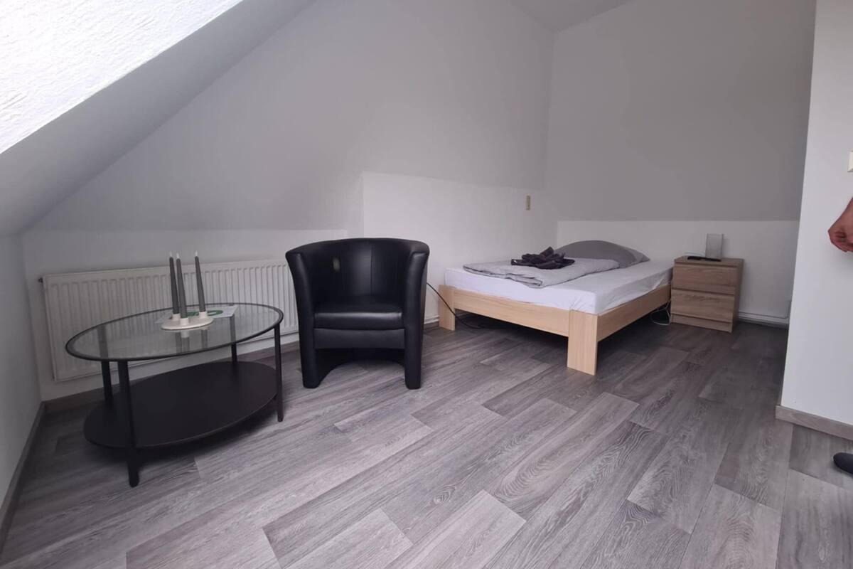 Un dormitorio pequeño y moderno con techo estilo loft, una cama con estructura de madera, un sillón de cuero negro y una mesa de vidrio con velas.