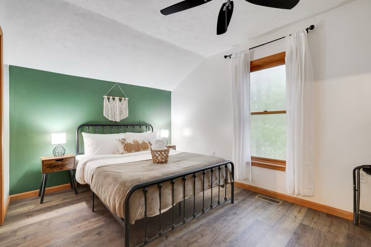 A bedroom with a bed, bedside tables, a ceiling fan, a window with curtains, and a green accent wall.