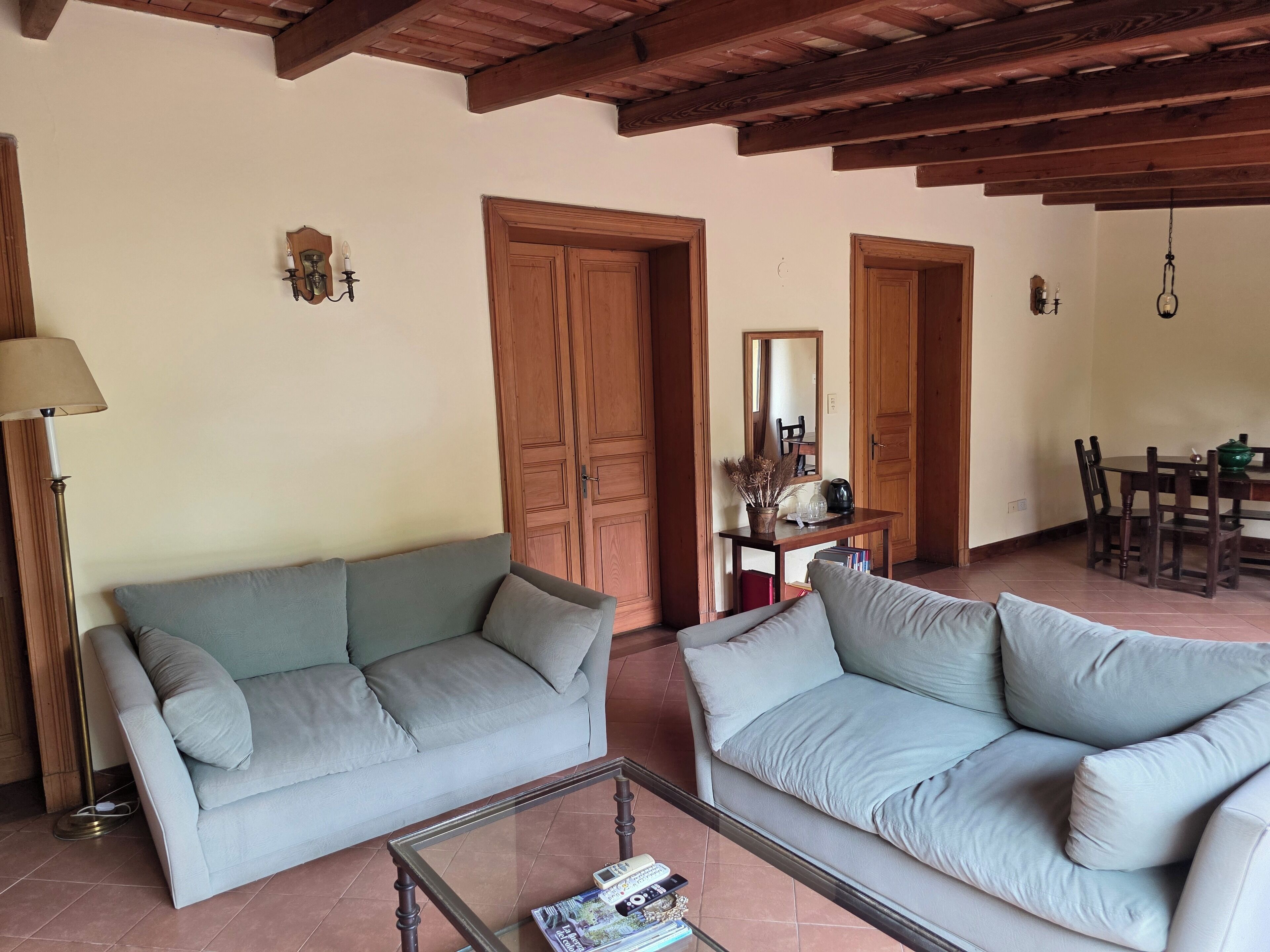 A living room with two light grey sofas, a glass coffee table, wooden doors, and a dining area with a table and chairs.