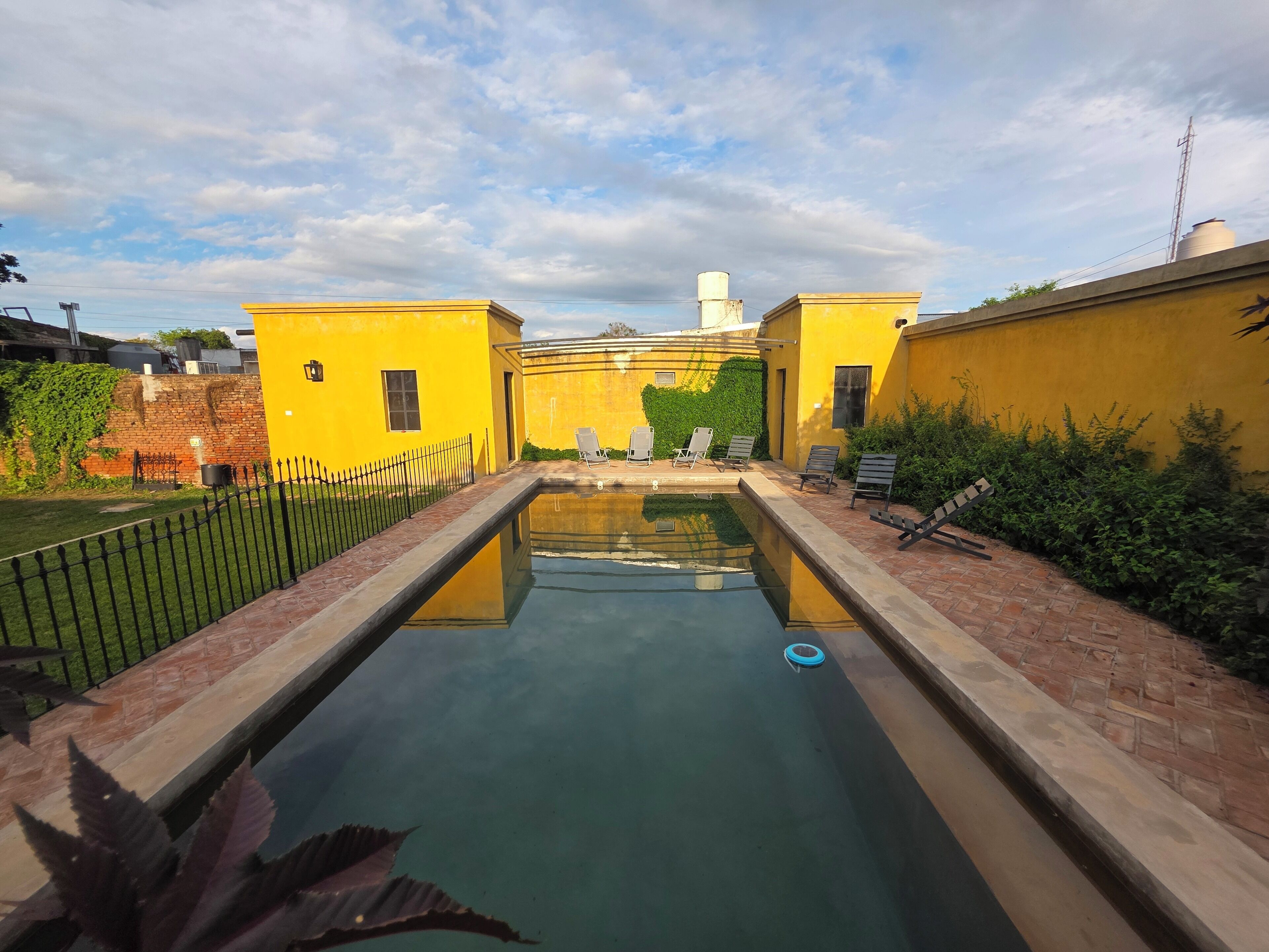 Una piscina con una vista despejada del cielo y un camino de ladrillo que lleva a un edificio amarillo.