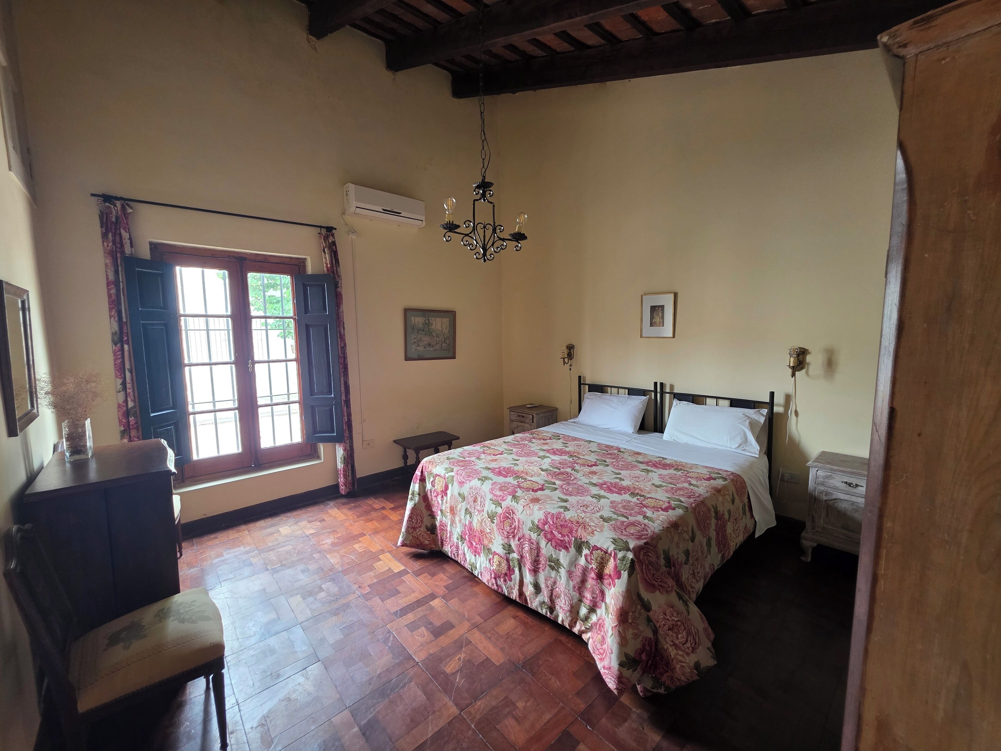 A bedroom with a bed, wooden floor, a window with curtains, a chandelier, and a nightstand.