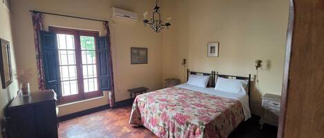 Un dormitorio con cama, suelo de madera, ventana con cortinas, candelabro y mesita de noche.