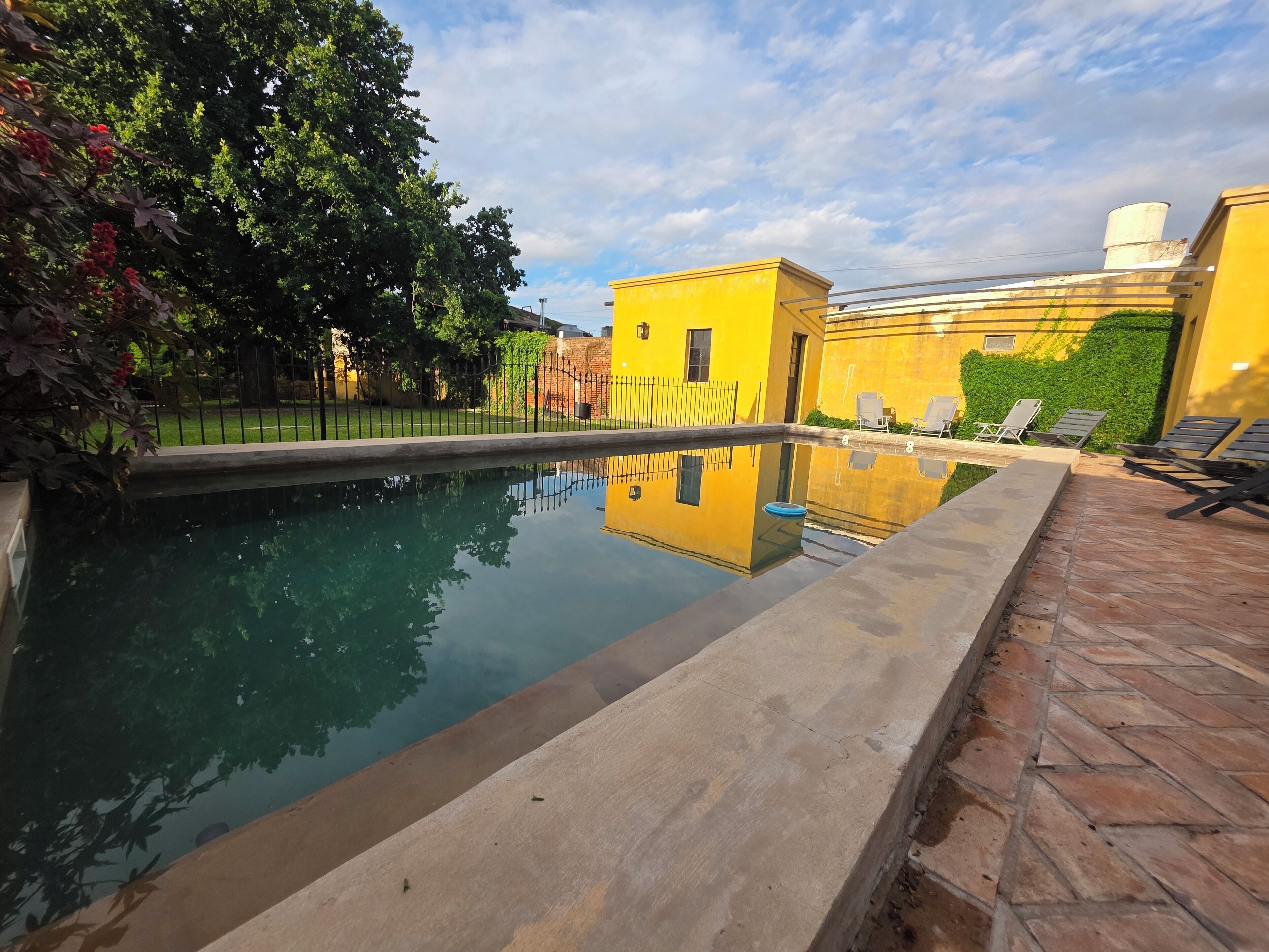 Una piscina con un riflesso limpido, circondata da un patio in mattoni e un edificio giallo con parete verde.