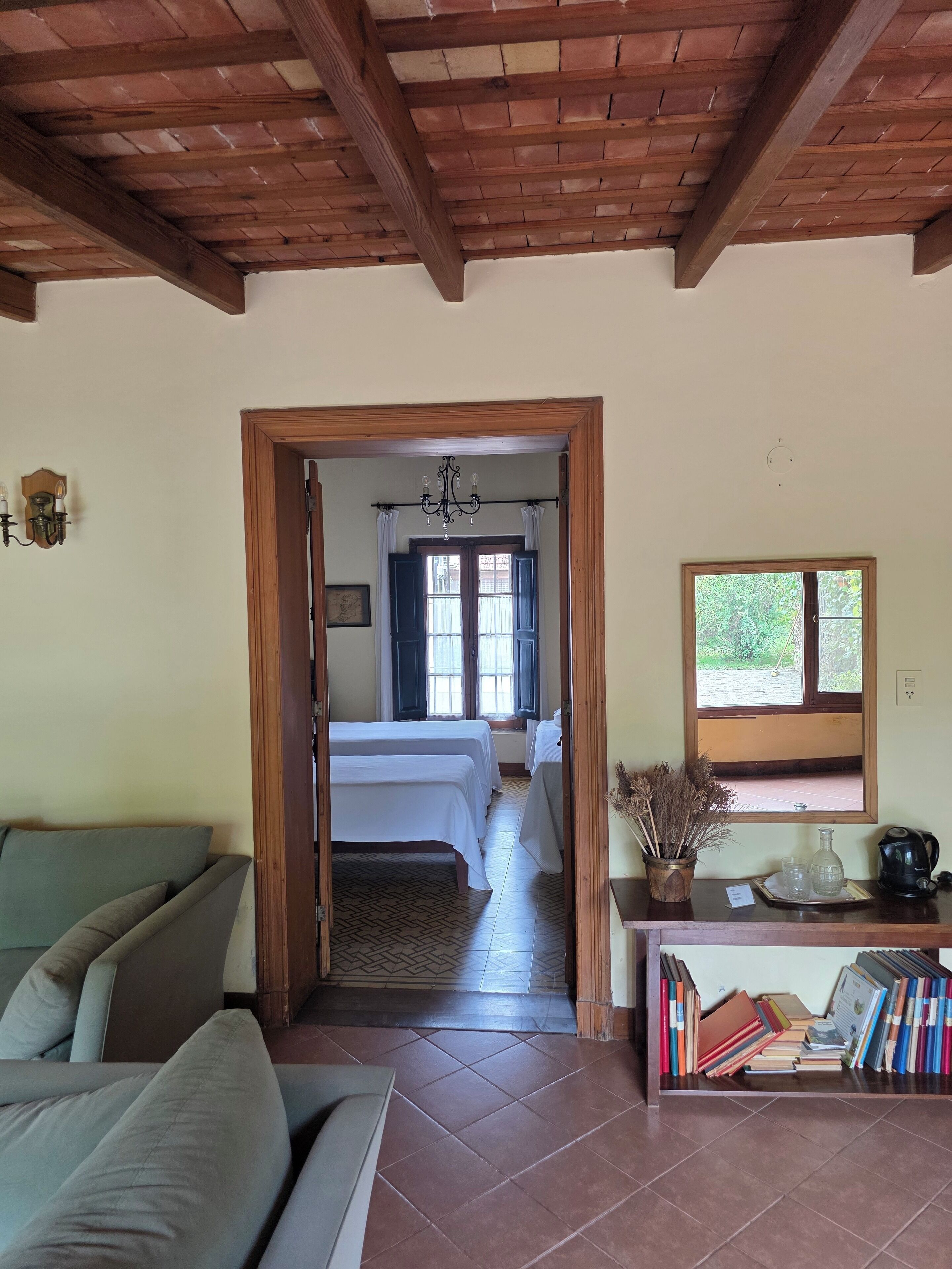 Una habitación con techo de madera, una cama con ropa blanca, un sofá, una mesita con libros y una tetera, y una ventana con vistas a zonas verdes.