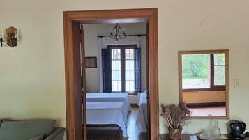 Una habitación con techo de madera, una cama con ropa blanca, un sofá, una mesita con libros y una tetera, y una ventana con vistas a zonas verdes.