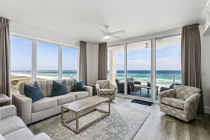 A spacious living room with a large window overlooking the beach, a ceiling fan, and a view of the ocean. - Newly Renovated 4th-Floor Gulf Front Condo – A Luxurious Coastal Retreat! Sleeps 10 (Navarre)