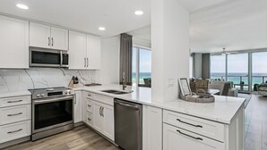 A modern kitchen with white cabinetry, stainless steel appliances, a large island with a sink, and a view of the ocean through large windows. - Newly Renovated 4th-Floor Gulf Front Condo – A Luxurious Coastal Retreat! Sleeps 10 (Navarre)