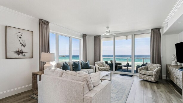 A spacious living room with a large sofa, armchairs, and a coffee table. There is a television on a wooden cabinet, and the room has large windows with a view of the ocean. - Newly Renovated 4th-Floor Gulf Front Condo – A Luxurious Coastal Retreat! Sleeps 10 (Navarre)