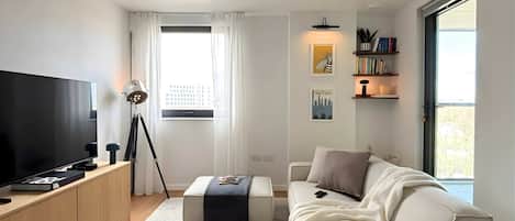 A modern living room with a flat-screen TV, a tripod, a bookshelf, a sofa, and a window with curtains.