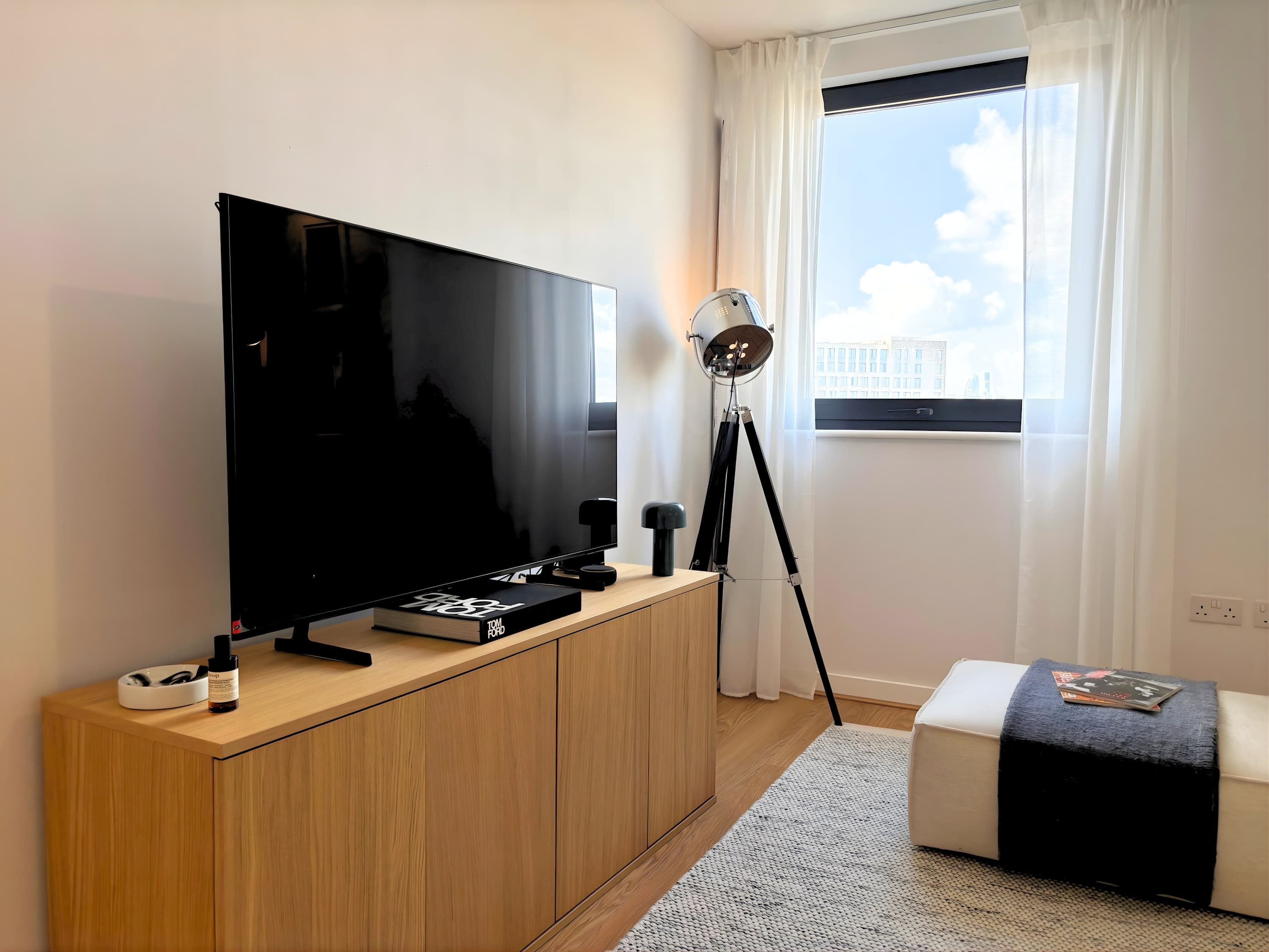 A modern living room with a flat-screen TV, a floor lamp, a small ottoman, and a window with a city view.