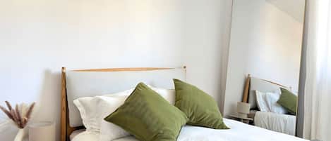 A neatly made bed with white bedding and green pillows, a bedside table with a lamp, and a large mirror on the wall.