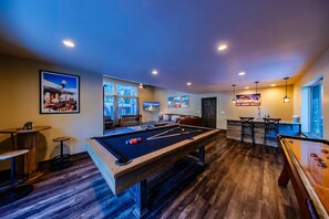 A pool table in a room with wooden flooring, a bar area with stools, and a large window providing a view of the outdoors. - Luxury Slopeside Ski Haus | Hot Tub + Sauna | 12P (South Lake Tahoe)