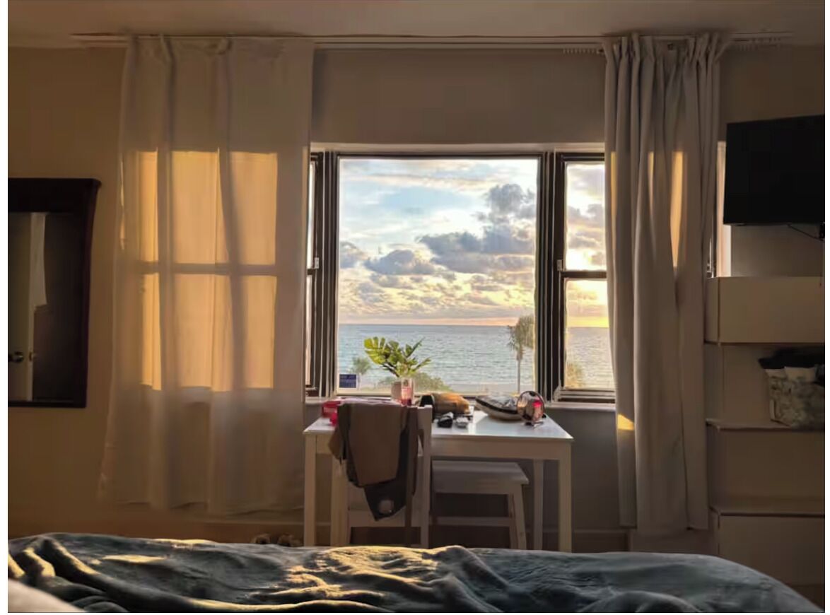 A room with a bed, a desk with a chair, a window with a view of the sea and clouds, and a TV on the wall.