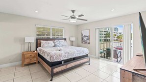 Una camera da letto con un letto, comodini, una ventola a soffitto, una finestra con persiane e un balcone con mobili da esterno, affacciata su edifici.