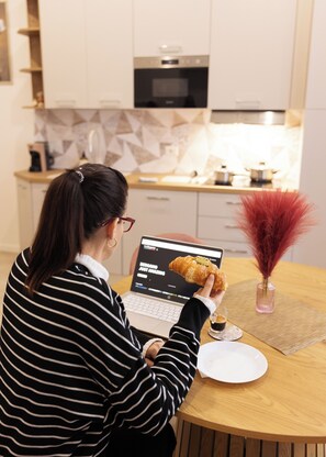 Eine Person sitzt an einem Holztisch mit einem Laptop, hält ein Croissant und eine Tasse Kaffee.