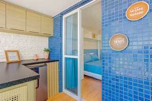 A compact hotel room with a kitchenette, a bed with blue bedding, and a bathroom with a glass door. - BR19 Beautiful Apartment 300m from Barra Beach (Salvador)