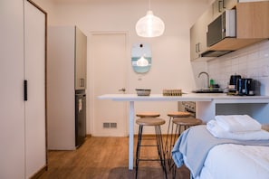 Eine kleine Küche mit weißem Arbeitsplatz, Barhocker, einer Pendelleuchte und einem Bett mit blauer Decke.