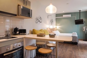 Eine kleine Küche mit Mikrowelle, Kaffeemaschine und Toaster. Ein Essbereich mit Hockern und ein Bett, im Hintergrund ist ein Fernseher zu sehen.
