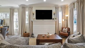 A living room with a flat-screen TV mounted on a white mantelpiece, a wooden coffee table, a brown leather sofa, and a grey armchair. - Beautifully Renovated Historic 4 BR Home|Sleeps 16 (Birmingham)