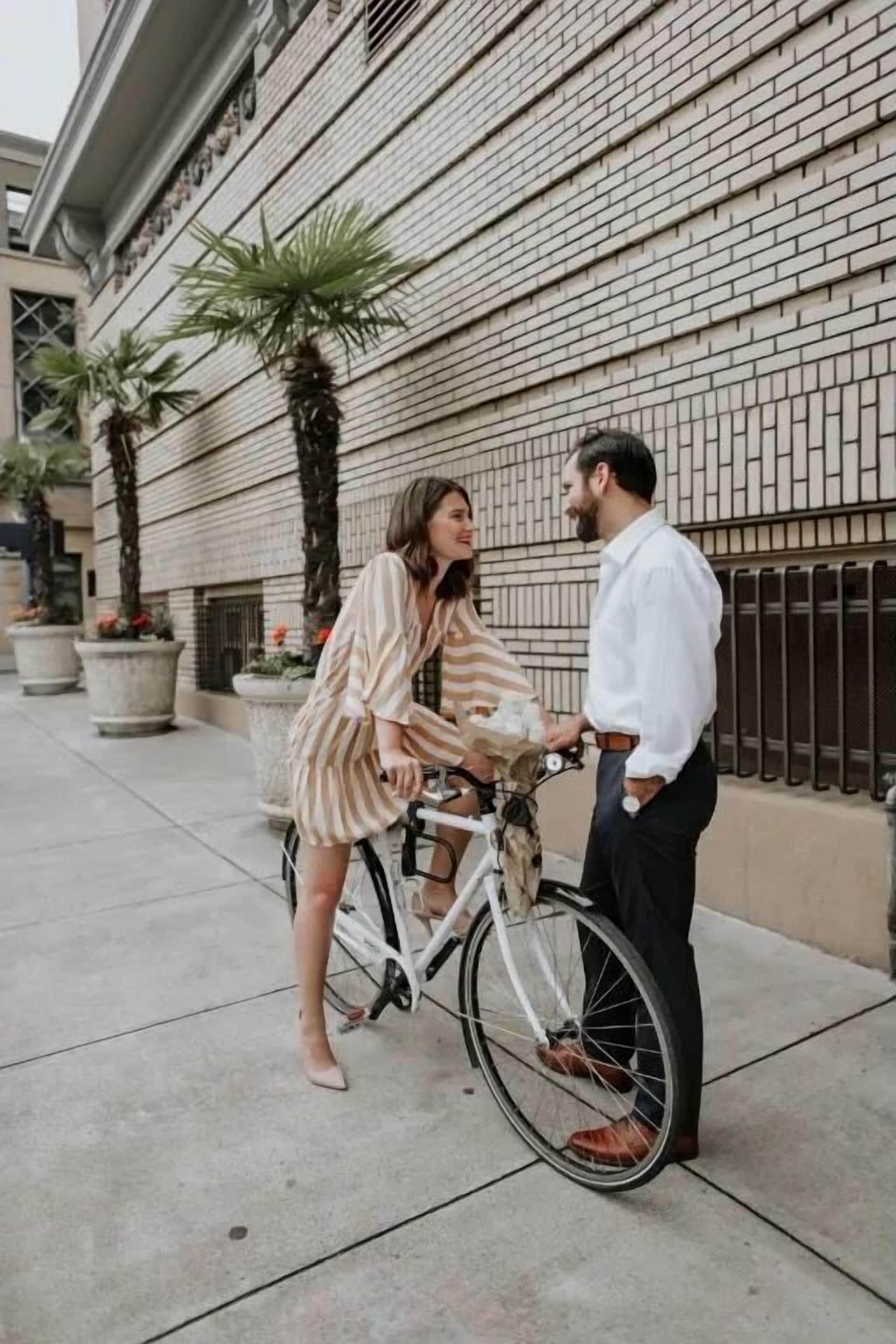 A person in a striped dress standing next to a white bicycle on a sidewalk.