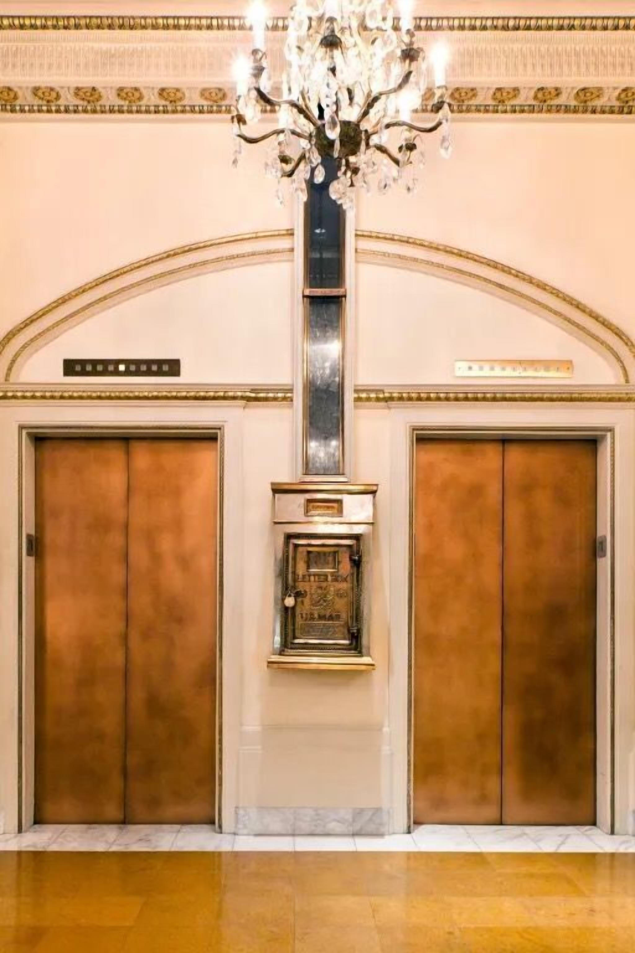 Una hall con due ascensori, un lampadario e una targa appesa al muro.