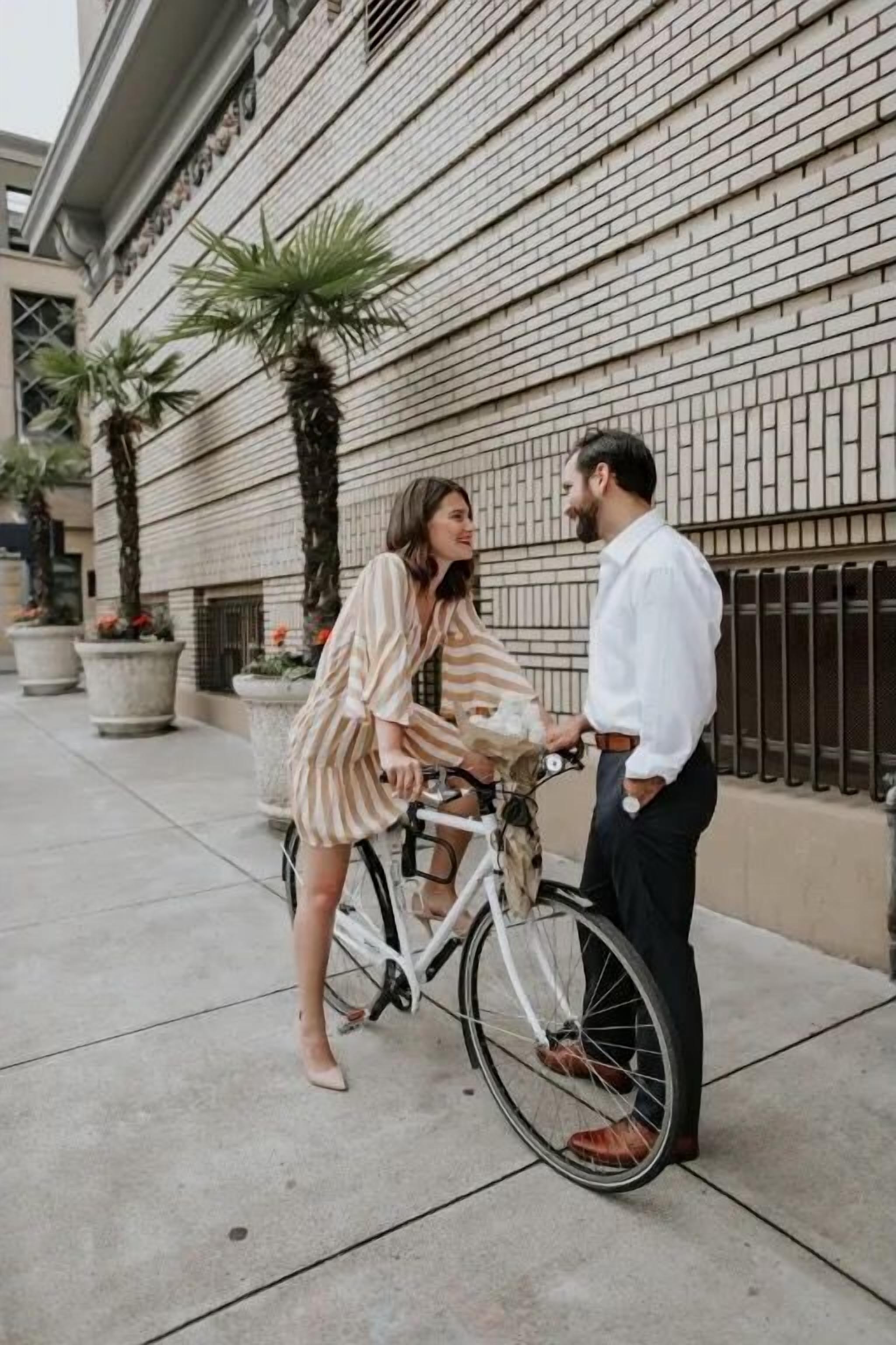 A person in a striped dress standing next to a white bicycle on a sidewalk.