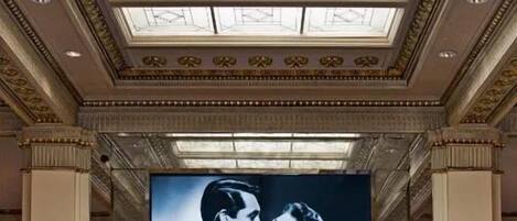 A lobby with a large digital screen displaying a black and white photo, columns, a round table with a vase of flowers, and a high ceiling with skylights.