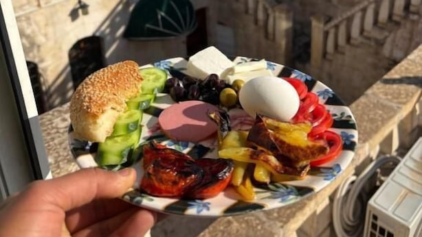 Un plato de comida con pan, queso, aceitunas, tomates y un huevo duro, sostenido por una mano.