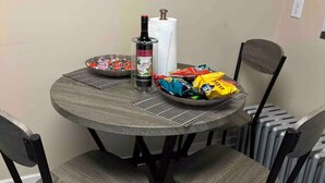 Una mesa redonda para comer con superficie de rejilla metálica, rodeada por cuatro sillas con patas oscuras y asientos acolchados. Hay una botella de vino y un rollo de servilletas sobre la mesa, junto con algunos aperitivos.
