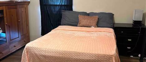 A bedroom with a bed, a dark-colored headboard, a wooden wardrobe, a small bedside table with a lamp, and a dark rug.