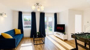 A modern living room with a blue sofa, a coffee table, a TV displaying 'NETFLIX', and a foosball table. - Stoke Mandeville House | Sleeps up to 6-8 (Aylesbury)