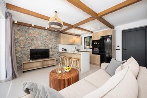 Una sala de estar moderna con pared de piedra, vigas de madera, un televisor, un sofá y una zona de comedor con taburetes.