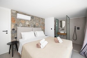Camera d'albergo con parete in pietra decorativa, un letto con lenzuola bianche, un comodino, un bagno con lavandino e specchio e una doccia.
