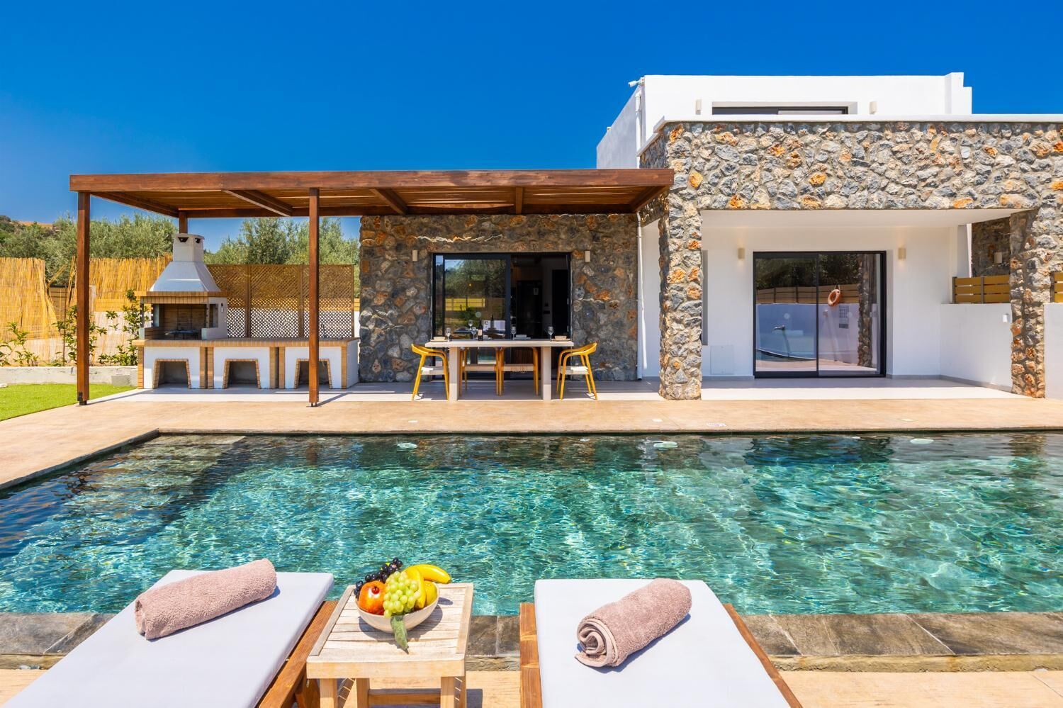 A modern outdoor pool area with a stone wall, a wooden pergola, and a dining table with chairs.