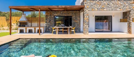 A modern outdoor pool area with a stone wall, a wooden pergola, and a dining table with chairs.